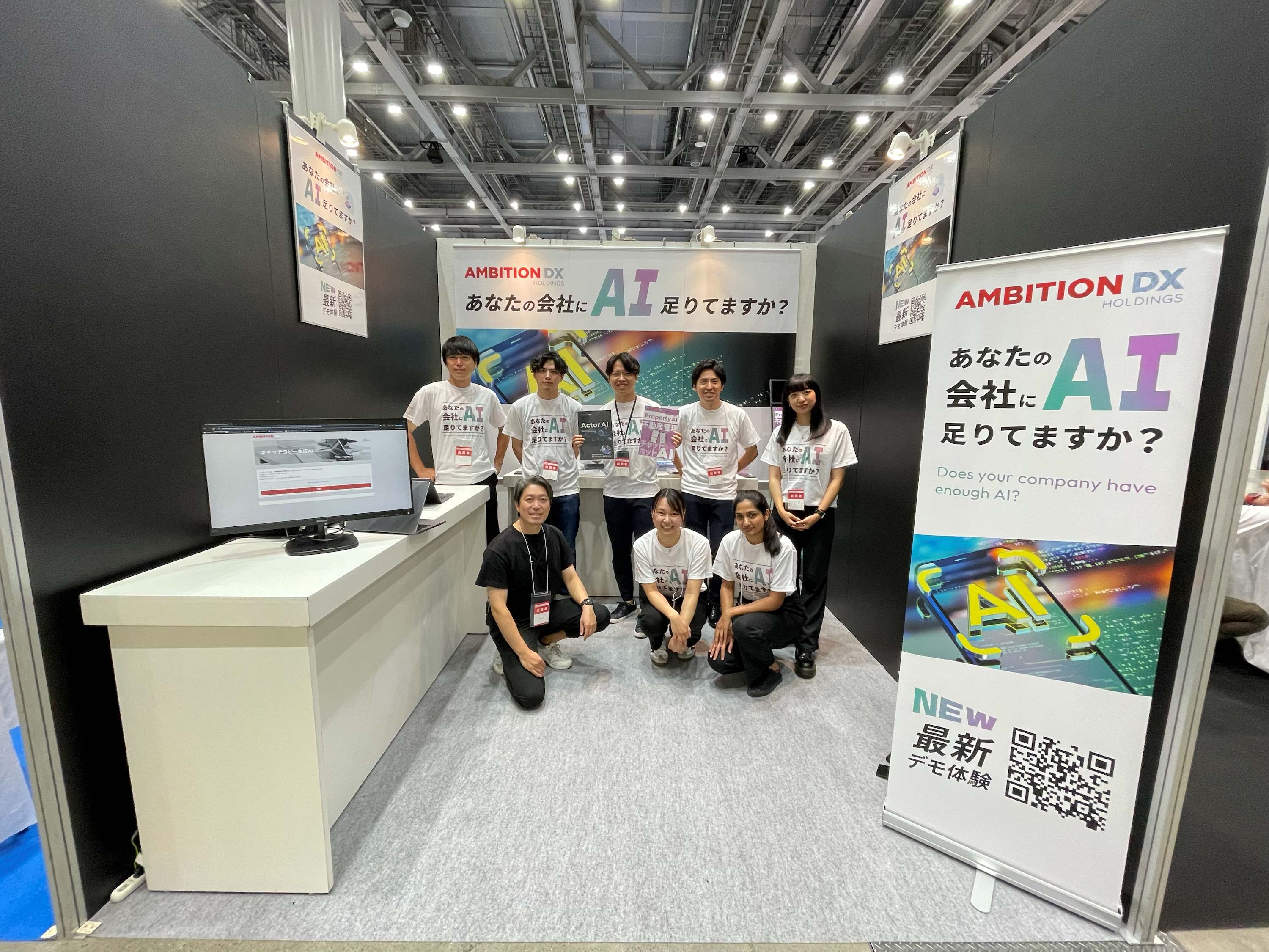 A group of eight team members from "AMBITION DX HOLDINGS" is posing at their trade show booth. They are wearing matching white t-shirts with "AI" prominently displayed, promoting their AI solutions.